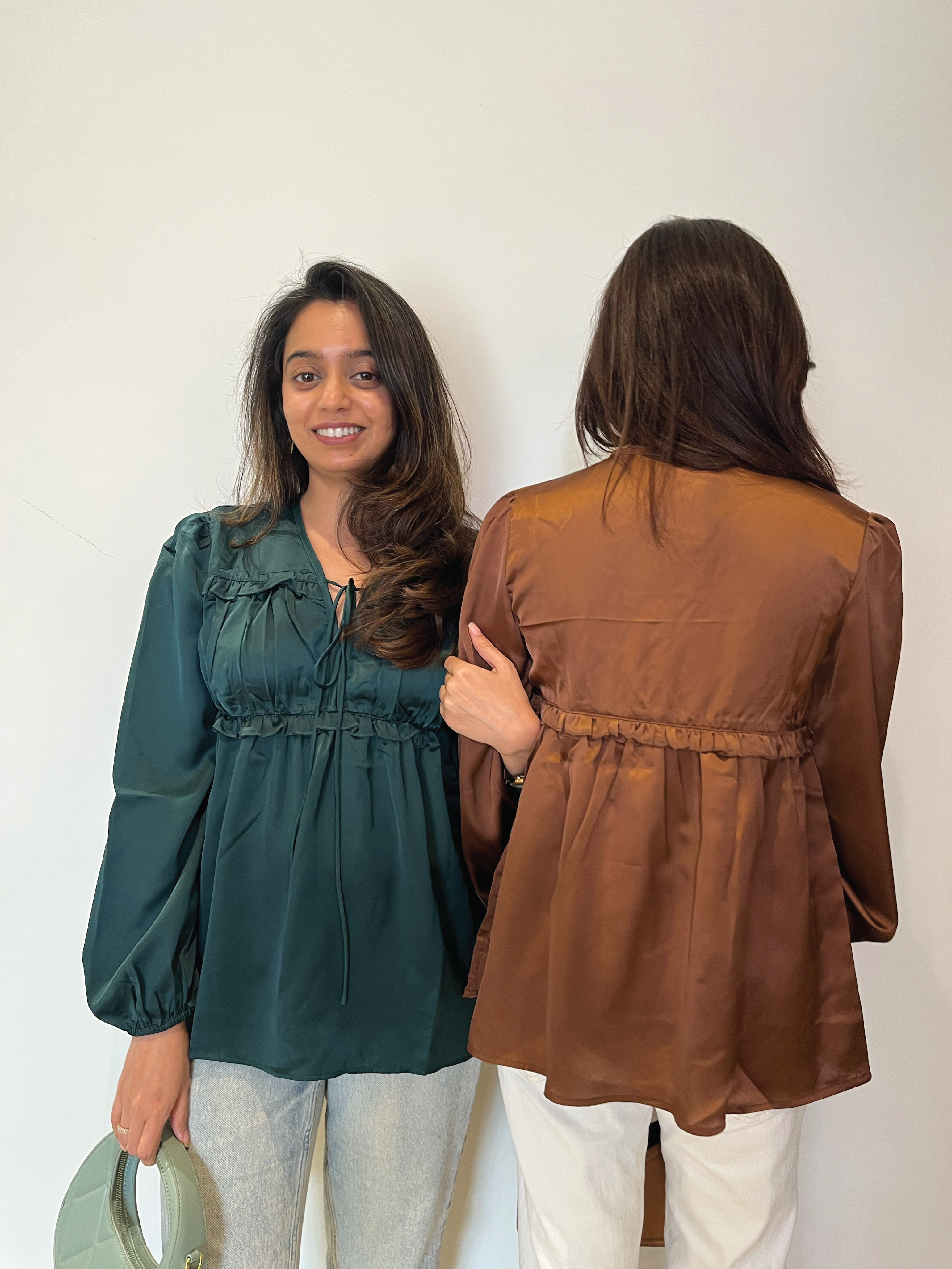 Two women wearing green and brown tops, standing back-to-back against a white wall wearing Rerasa Satin Pleated Tie Up Top