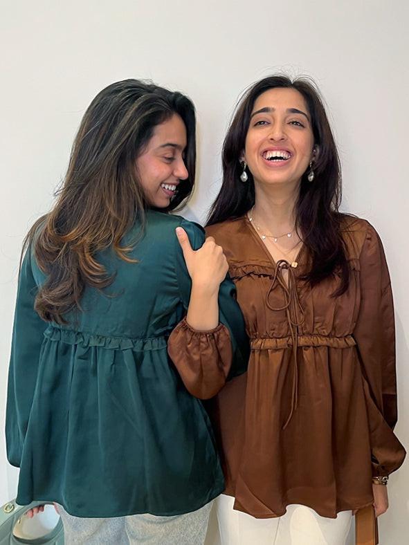 Two women wearing green and brown tops, smiling and interacting with each other against a plain background. Satin pleated tie up top